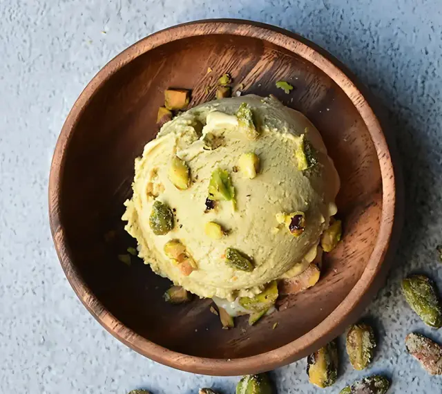 Cremoso helado de pistacho servido en un cuenco de madera, adornado con trozos de pistacho y acompañado de más pistachos enteros esparcidos alrededor.